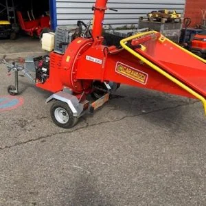 Un broyeur de bois Broyeur de Branches rouge avec un garde-corps jaune est garé sur une aire pavée à l'extérieur d'un garage aux portes bleues.