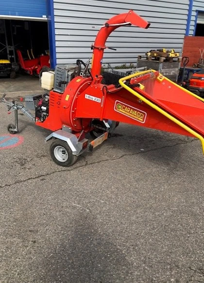 Un broyeur de bois Broyeur de Branches rouge avec un garde-corps jaune est garé sur une aire pavée à l'extérieur d'un garage aux portes bleues.